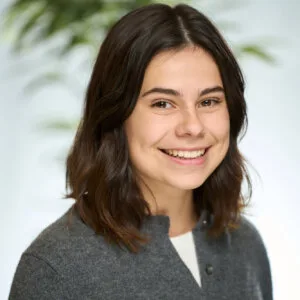 A woman, Tiana Alphonso, has shoulder length dark brown hair and wears a grey buttoned sweater. She smiles warmly.