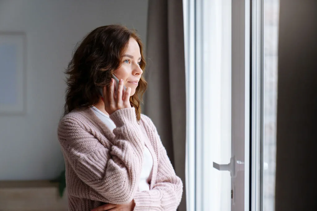 Young woman talking on mobile phone at home, looking through a window.