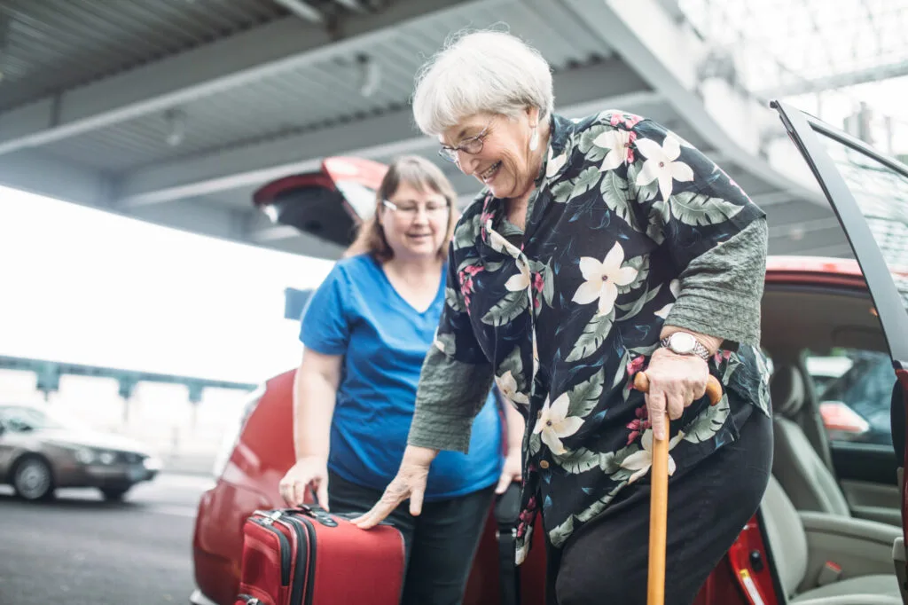 A mature woman in her 70's gets out of a car while holding a cane with a female friend at the airport. The friend hands the woman her luggage. Both women have a cheerful smile on their faces.